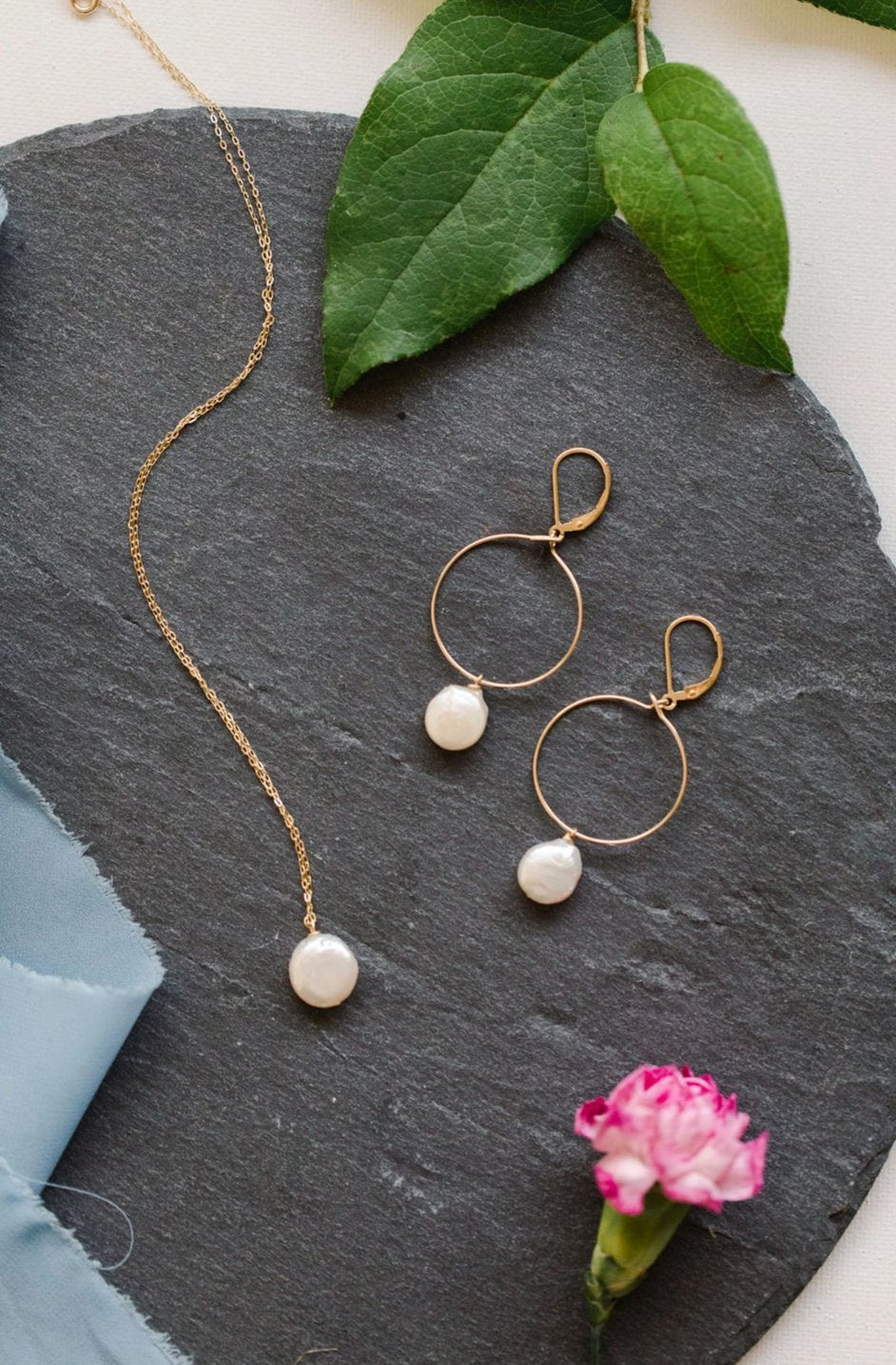 Gold and freshwater coin pearl necklace and hoop earring set on a grey slate backdrop with green leaves, a pink carnation, and blue silk ribbon styling