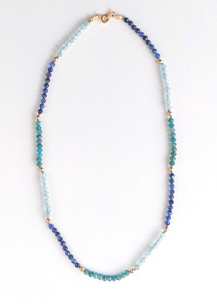 Portrait cropped image of a blue ombre beaded gemstone necklace with tiny beads of aquamarine, kyanite, and apatite. Gemstone beads strung on silk thread with gold accents beads and lobster clasp. Handcrafted jewelry by artist