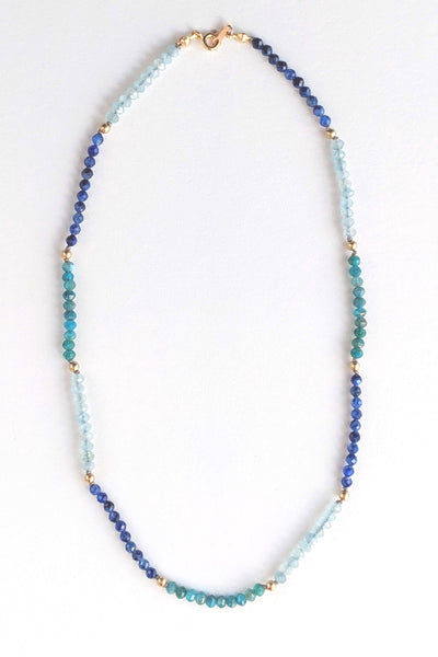 Portrait cropped image of a blue ombre beaded gemstone necklace with tiny beads of aquamarine, kyanite, and apatite. Gemstone beads strung on silk thread with gold accents beads and lobster clasp. Handcrafted jewelry by artist