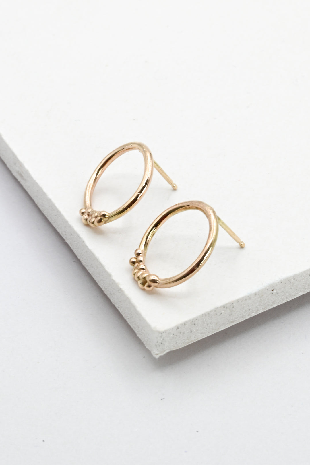 A pair of gold hoop post earrings with small textured bead details at the front, displayed on a white textured rectangular block against a clean white background.