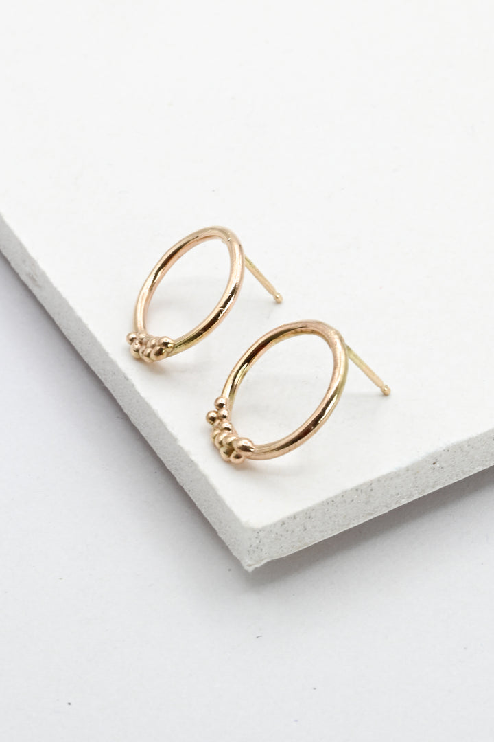 A pair of gold hoop post earrings with small textured bead details at the front, displayed on a white textured rectangular block against a clean white background.
