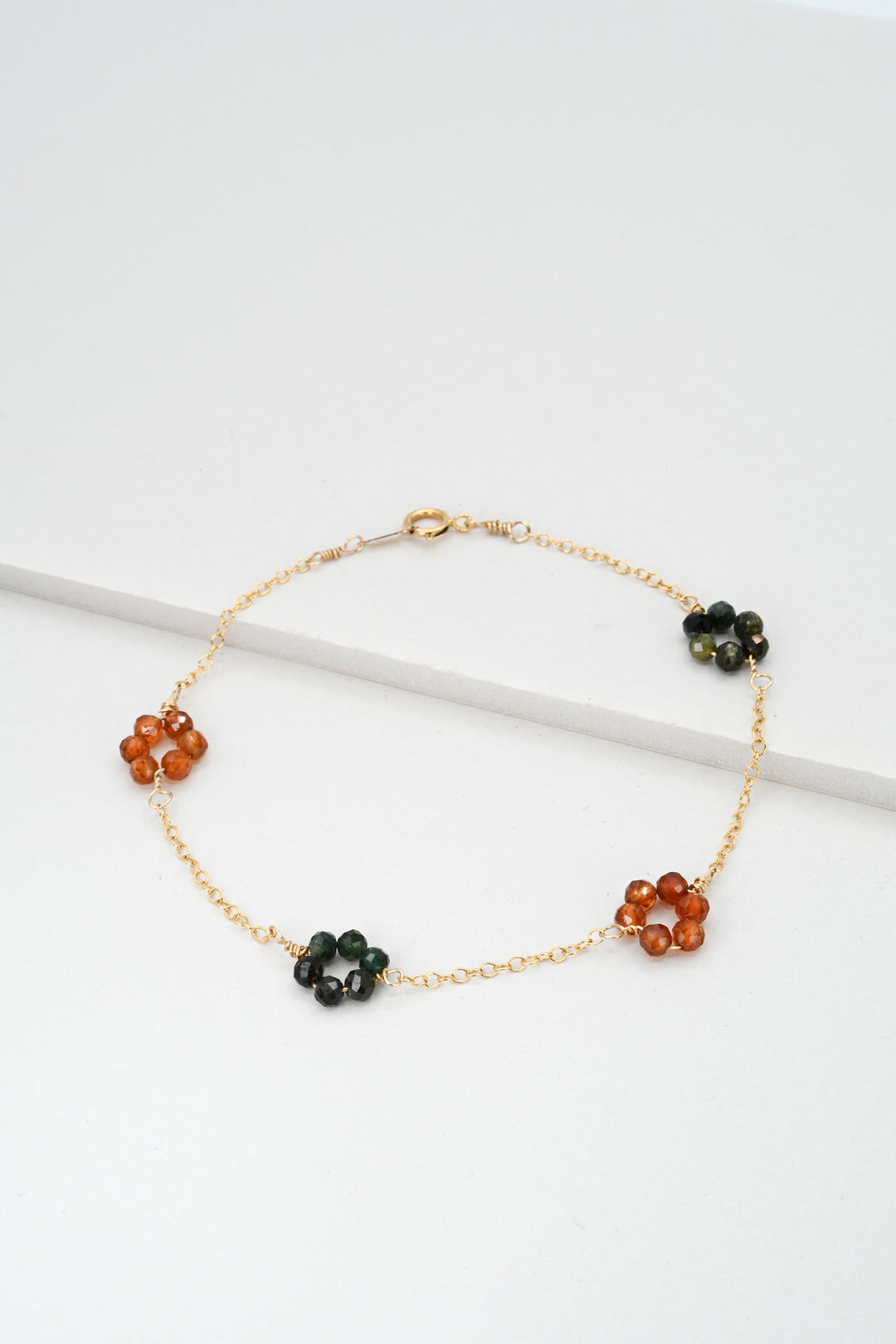 Gold daisy chain bracelet with green tourmaline and orange hessonite garnet "flowers" on a white background.