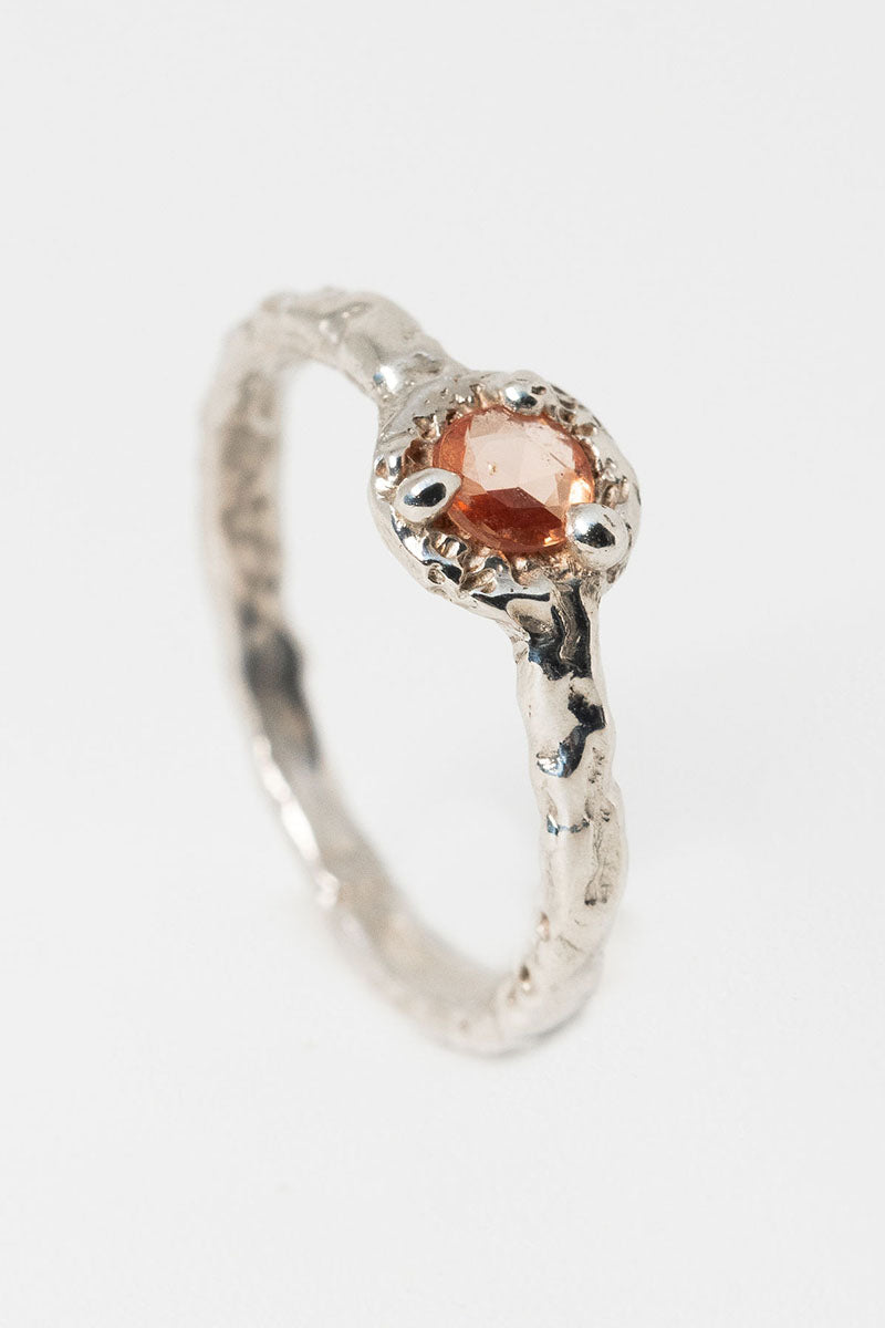 A textured silver ring with peach sapphire solitaire stone set at an angle on a white background