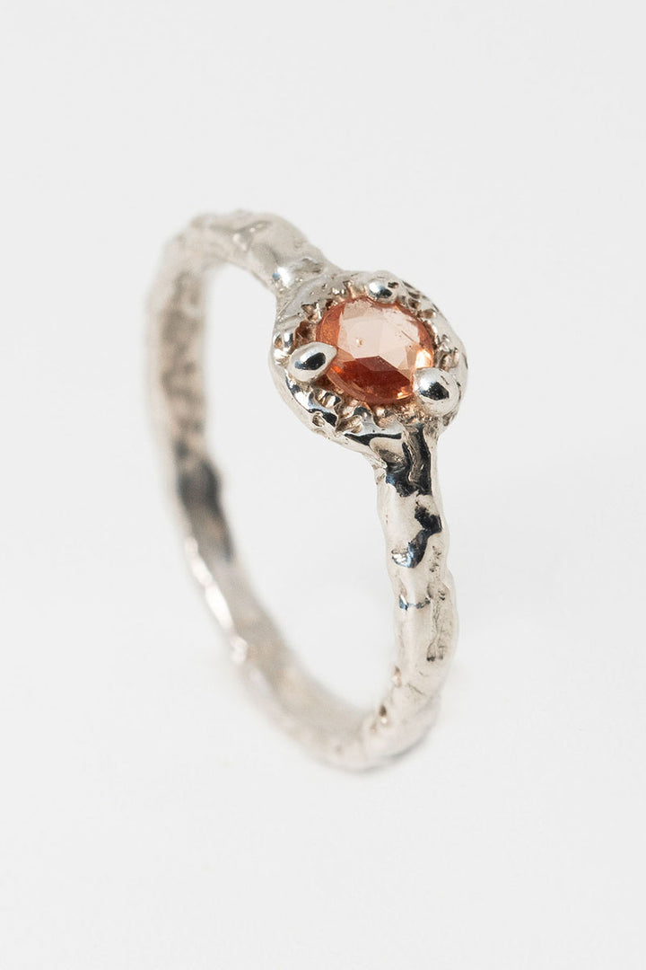 A textured silver ring with peach sapphire solitaire stone set at an angle on a white background