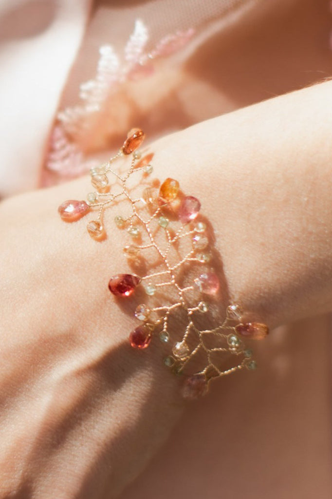 A watermelon tourmaline and peridot bracelet in the form of a delicate gold vine by J'Adorn Designs is modeled on the wrist of a white woman wearing a pink slip