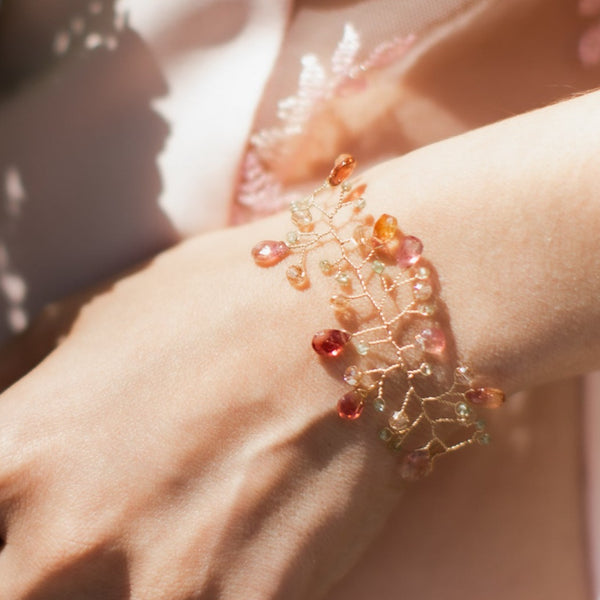 A watermelon tourmaline and peridot bracelet in the form of a delicate gold vine by J'Adorn Designs is modeled on the wrist of a white woman wearing a pink slip