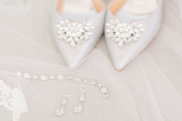 Flat lay photo of sparkly blue and crystal wedding accessories, blue satin shoes, double teardrop silver bridal earrings, and pear shaped crystal wedding bracelet jewelry set. 