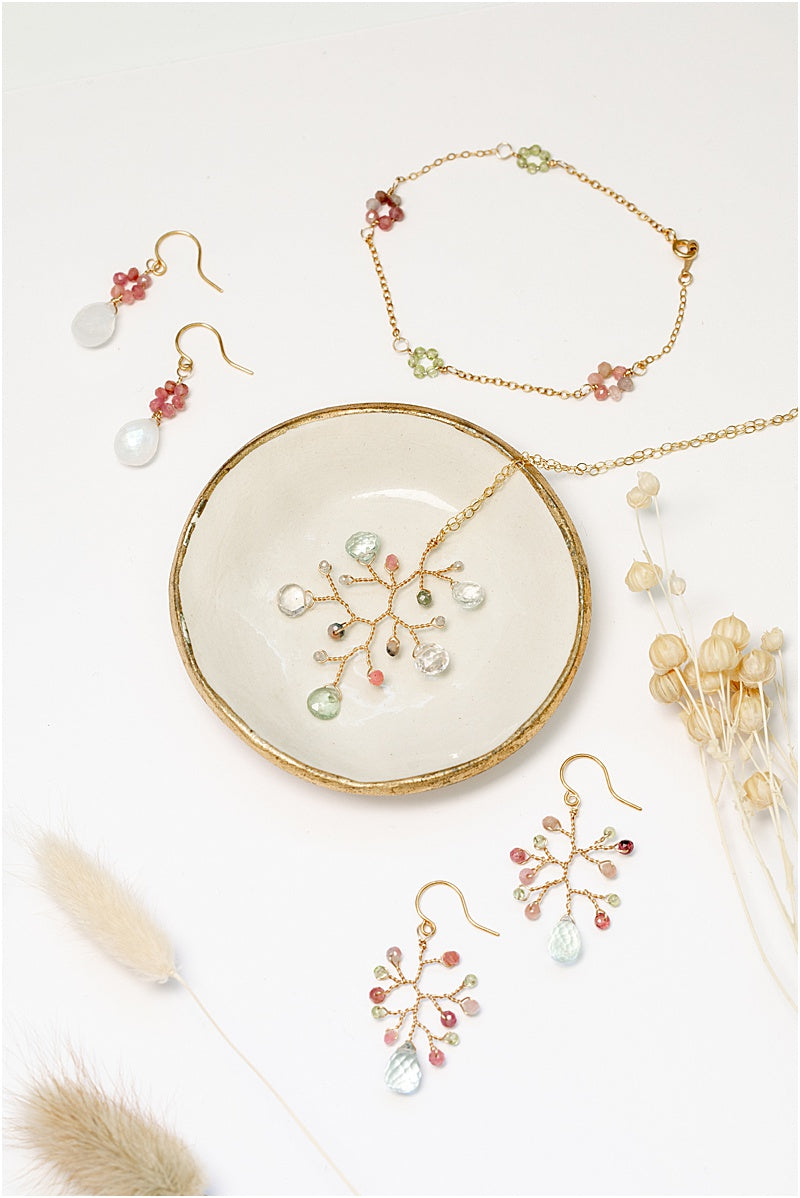 A matching gold jewelry collection with colorful gemstone flowers. Handcrafted jewelry set features a necklace, a bracelet, and two pairs of earrings on a gold-rimmed ivory ceramic dish. For sale: one handcrafted gold pendant featuring a wire-wrapped branch with aquamarine teardrops and tourmaline beads in a rainbow of colors. A lightweight, hypoallergenic gemstone necklace on a fine 14k gold filled chain. Artisan jewelry made by Alison Jefferies for J'Adorn Designs.