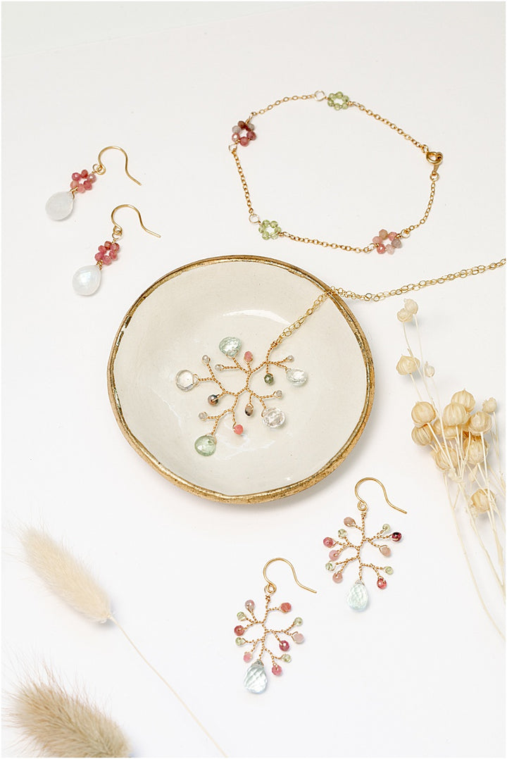 A matching gold jewelry collection with colorful gemstone flowers. Handcrafted jewelry set features a necklace, a bracelet, and two pairs of earrings on a gold-rimmed ivory ceramic dish. For sale: one handcrafted gold pendant featuring a wire-wrapped branch with aquamarine teardrops and tourmaline beads in a rainbow of colors. A lightweight, hypoallergenic gemstone necklace on a fine 14k gold filled chain. Artisan jewelry made by Alison Jefferies for J'Adorn Designs.